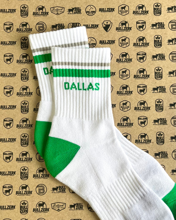 A pair of white crew socks with green and gray stripes and 'DALLAS' text laying on a patterned background. 
