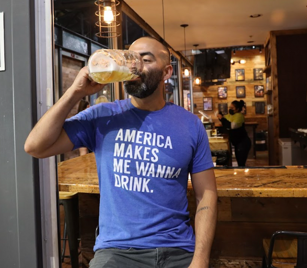 Photo of a man sitting at a bar counter, drinking a beer and wearing the Bullzerk America Makes Me Wanna Drink tshirt. 