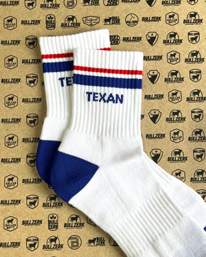 A pair of white crew socks with blue and red stripes and 'TEXAN' text laying on a patterned background. 