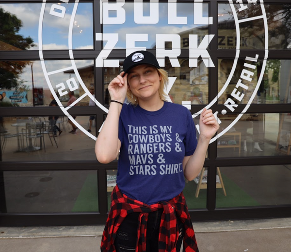 Woman wearing a Bullzerk This is my Cowboys, Rangers, Mavs and Stars tshirt and a baseball hat, standing in front of a glass garage door. 