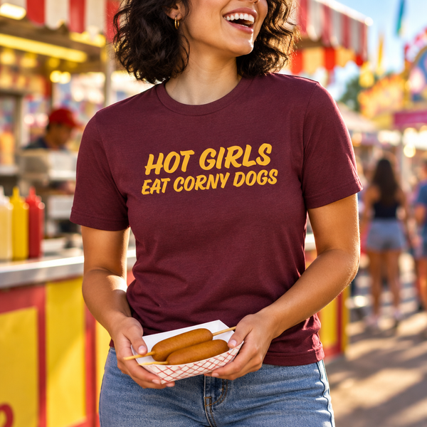 Woman wearing maroon Hot Girls Eat Corny Dogs T-shirt at a fair food stand, holding corny dogs