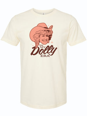 A cream shirt with a light orange and maroon illustration of Dolly Parton wearing a cowboy hat that reads 