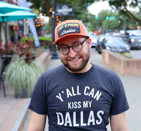 Man wearing a Bullzerk Y'all Can Kiss My Dallas Tshirt and Dallas Skyline patched hat, standing on the sidewalk of a busy street. 