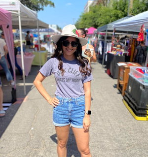 Woman at an outdoor pop-up market, wearing sunglasses, a hat and a Bullzerk  Oak Cliff that's my Hood tshirt. 