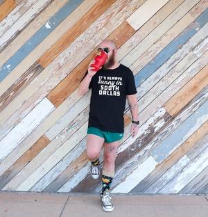 Man leaning against a wooden wall, wearing a Bullzerk Sunny in South Dallas tshirt and drinking from a red cup. 