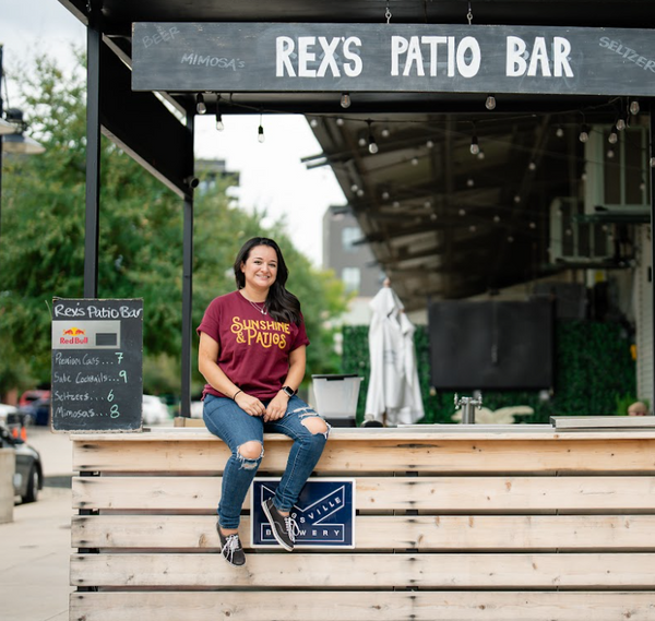 Woman sitting on a wooden bench at Rex's Patio Bar, wearing a Bullzerk Sunshine and Patios tshirt. 