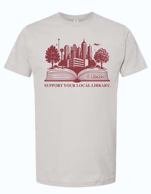 Light gray unisex t-shirt featuring the design 'Support Your Local Library' with a skyline illustration representing Dallas. Perfect for showing love for literature and local community, this shirt is a great Texas themed gift, Texas Made Gifts, and a souvenir from Texas for book lovers or as a Dallas souvenir. Ideal for those looking for unique Texan gifts.