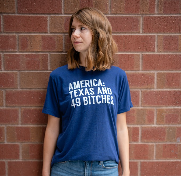 Person wearing a navy t-shirt with text "America: Texas and 49 Bitches" standing against a brick wall.