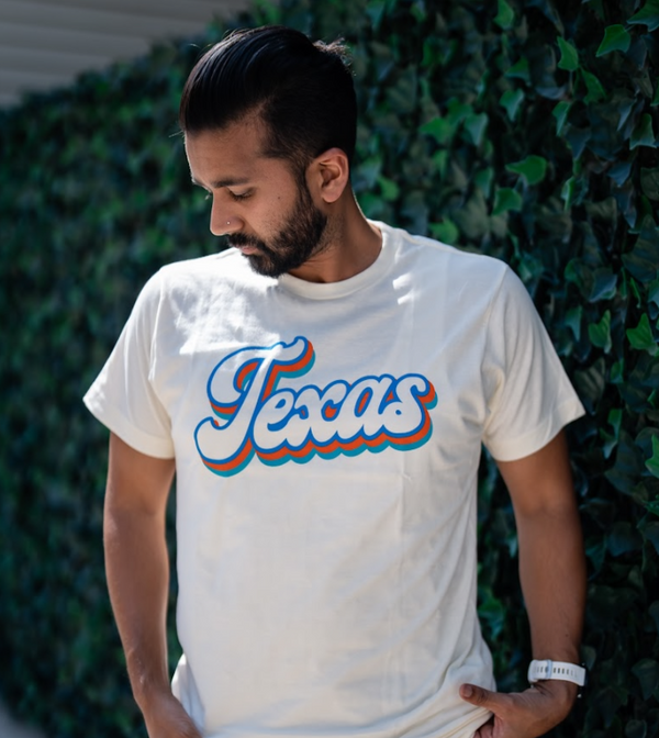 Man wearing a cream t-shirt with 'Texas' printed on it in a script font, standing against a green ivy wall.