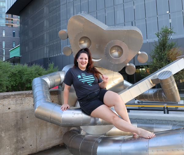 Woman sitting on a large metal statue of a robot, wearing a Bullzerk Texas Silhouette tshirt. 