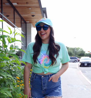 Woman wearing the Bullzerk Tie Dye Texas Silhouette tshirt with sunglasses and a baseball hat, standing outside near flowers. 