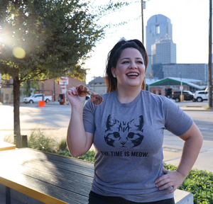Woman standing outside downtown Dallas, wearing a Bullzerk Time is Meow tshirt and holding sunglasses. 