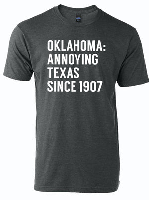 A dark gray unisex fine jersey t-shirt featuring the text 'Oklahoma: Annoying Texas Since 1907'. Perfect for those looking for a Texas themed gift, this stylish tee makes a great addition to any collection of Texas Made Gifts or a fun souvenir from Texas, especially in Dallas. Ideal for anyone searching for unique Texan gifts.