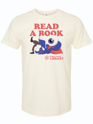 A playful illustration of a blue character reading a book on a beige unisex t-shirt featuring the title 'Read A Book.' This Texas themed gift supports the Dallas Public Library and makes for a great souvenir from Texas, perfect for book lovers and those looking for Texan gifts.